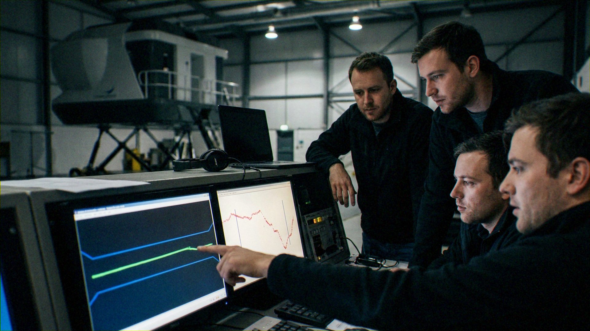 ['A team of four technicians or engineers', 'Computer-based data acquisition systems', 'Large-scale mechanical hardware (visible in background)']. Collaborative technical analysis where one individual directs the group's attention to specific performance metrics. Aerospace.