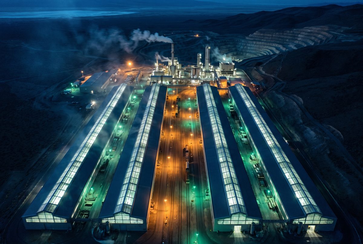 A vast munitions manufacturing campus at night from the air: four parallel production halls glowing from within, a minerals processing plant, and a worked extraction terrace stepping down into the hillside.