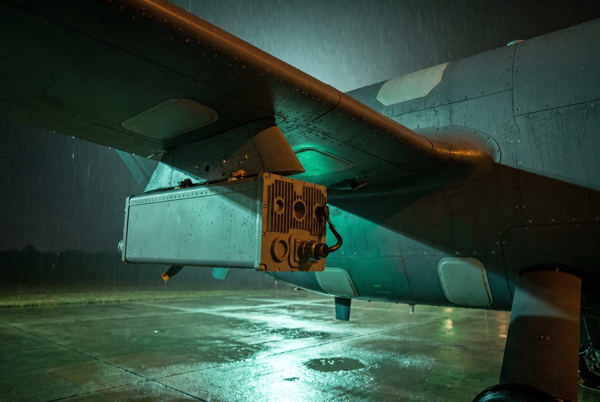 A sealed matte grey line-replaceable unit with milled cooling fins mounted on the wing pylon of a large uncrewed aircraft, photographed from below at night on a wet hardstand under emerald floodlight.